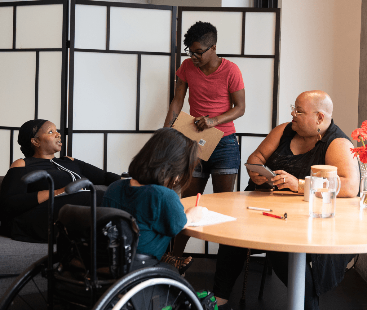 Cuatro personas discapacitadas de color están en torno a una mesa durante una reunión. Una mujer negra sentada en un sofá habla con una expresión neutral mientras las otras tres personas escuchan (una persona del sur de Asia que toma notas sentada en una silla de ruedas; una persona negra no binaria sentada en una silla, que sostiene una tableta y tiene un bastón, y una persona negra no binaria que está de pie y tiene un portapapeles en la mano).