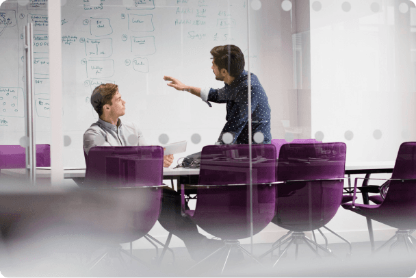 Two men in a modern meeting room, one gesturing at a whiteboard while the other listens.
